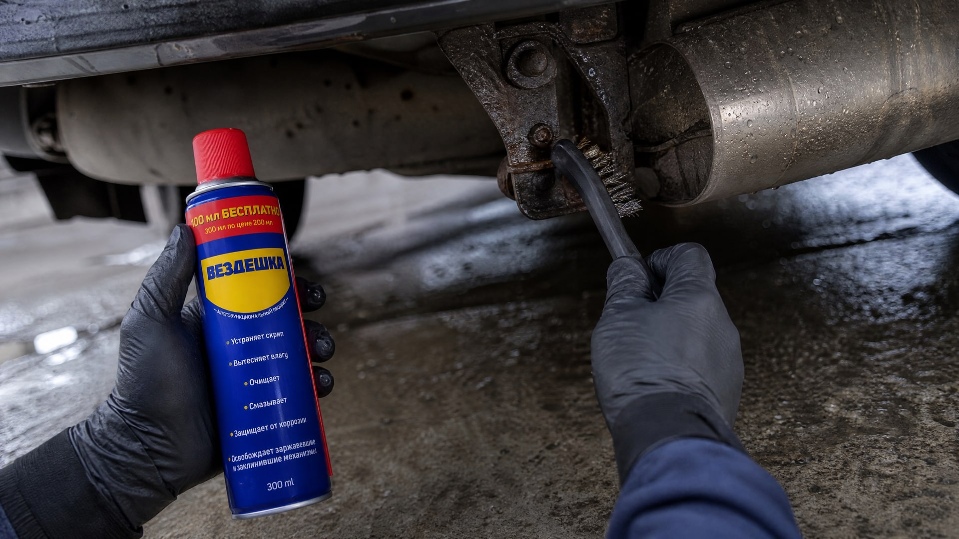 Рука в перчатке наносит WD-40 на болт глушителя