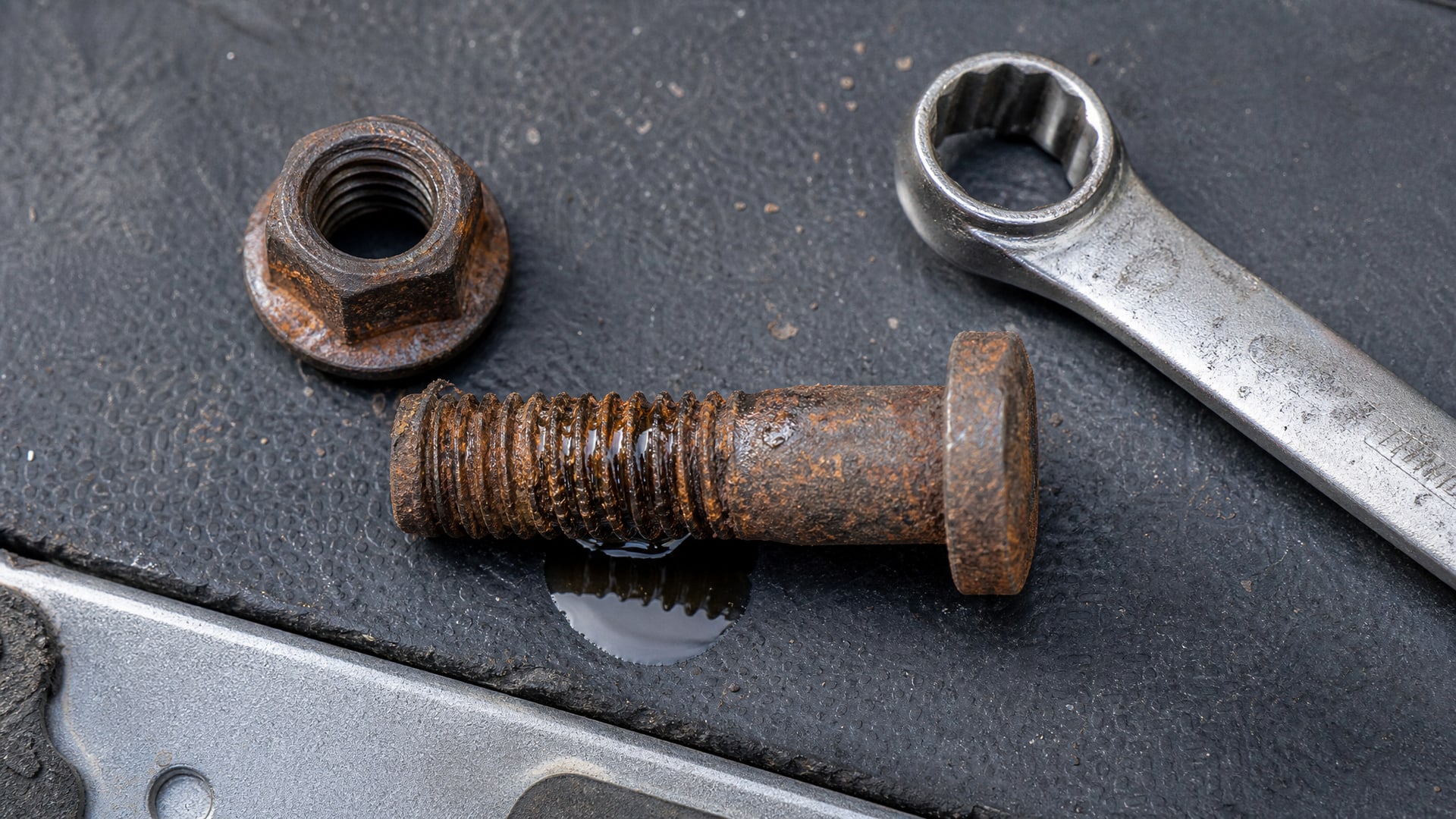 На верстаке лежат ржавый болт с гайкой, вокруг них видна лужица WD40.